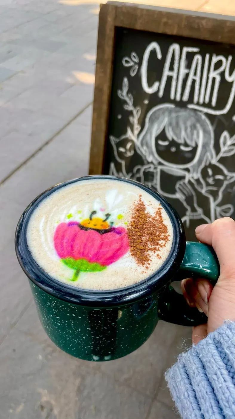 Taza de peltre verde con una flor rosa impresa, frente a una pizarra con el nombre Cafairy